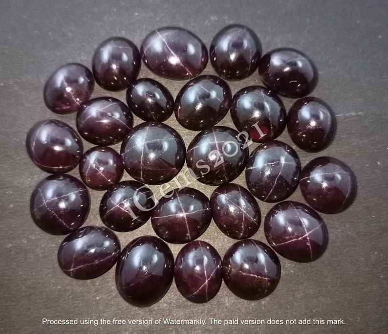May include: A collection of 20 dark red star-shaped gemstones, also known as star garnets, arranged in a circle on a dark brown surface. The gemstones are oval-shaped and have a star-like pattern in the centre.