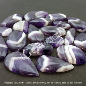 K&ouml;nnte beinhalten: Eine Sammlung von 20 amethystfarbenen Cabochons in Tropfenform mit violetten und wei&szlig;en Streifen.