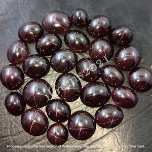May include: A collection of 25 dark red star-shaped gemstones, also known as star garnets, arranged in a circle on a dark grey surface.
