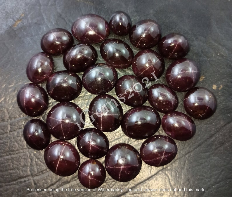 May include: A collection of 25 dark red star-shaped gemstones, also known as star garnets, arranged in a circle on a dark grey surface.