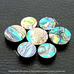 Könnte beinhalten: Sieben regenbogenfarbene runde Abalone-Muschel-Cabochons in verschiedenen Blautönen, Grüntönen und Goldtönen. Die Cabochons sind in einem Kreis auf einer schwarzen Oberfläche angeordnet.