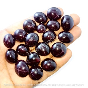 May include: A collection of 16 dark red, oval-shaped gemstones with a star-like pattern inside. The gemstones are arranged in a hand.