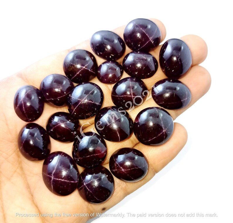 May include: A collection of 16 dark red, oval-shaped gemstones with a star-like pattern inside. The gemstones are arranged in a hand.