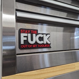 May include: A red and white sticker with the text "Stay the Fuck Out of My Toolbox" on a silver metal toolbox.
