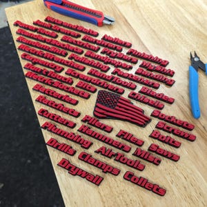 May include: A wooden surface with red and black labels for various tool categories. The labels include "Wallers", "Finishing", "My Stuff", "Junk Tools", "Electrical", "Sandpaper", "Fasteners", "Hand Tools", "Blades", "Chisels", "Sockets", "Screws", "Bolts", "Saws", "Files", "Pliers", "Hammers", "Cutters", "Ratchets", "Batteries", "Metric Sockets", "Don't Touch", "Carpentry", "Screwdrivers", "Random Shit", "Standard Sockets", "Power Tools", "Drywall", "Clamps", "Air Tools", "Drills", "Plumbing", and "Collets".