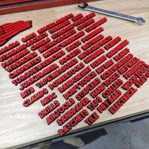 May include: Red plastic labels with the names of various tools and hardware items, including "Nailers", "Saws", "Miscellaneous", "Standard Sockets", "Hammers", "Plumbing", "Chisels", "Standard Wrenches", "Metric Sockets", "Cutters", "Carpentry", "Metric Wrenches", "Fasteners", "Torque Wrenches", "Bolts", "Polishing", "Junk Drawer", "Sockets", "Batteries", "Bits", "Tape Measures", "Nuts and Bolts", "Clamps", "Power Tools", "Sanding Screws", "Misc", "Random Shit", "Pliers", "Hardware", "Drills", "Don't Touch My Shit", "Blades", "Air Tools", "Staplers", "Screwdrivers", "Sandpaper", "Hand Tools", "Drywall", "Ratchets", "Taps", "Collets", "Finishing", "Tile", "Pry Bars", "Wood Tools", and "Electrical Stay Out".