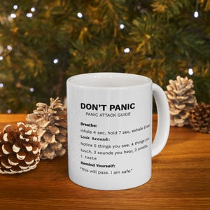 Puede incluir: Taza de cerámica blanca con el texto "DON'T PANIC" y una guía para ataques de pánico. La guía incluye instrucciones para respirar, mirar alrededor y recordarte a ti mismo que estás a salvo. La taza está sobre una superficie de madera.