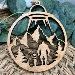May include: A wooden ornament featuring a silhouette of a Bigfoot and an alien standing in a forest with a UFO hovering above. The ornament is a light brown color and has a circular shape.
