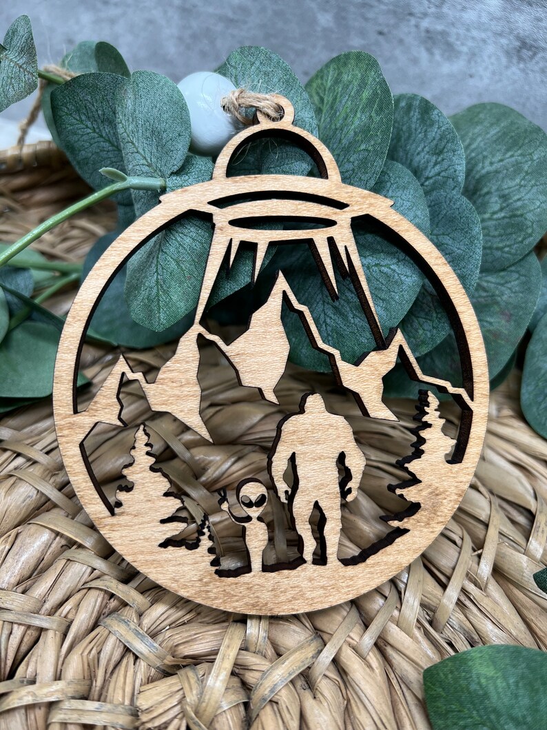 May include: A wooden ornament featuring a silhouette of a Bigfoot and an alien standing in a forest with a UFO hovering above. The ornament is a light brown color and has a circular shape.