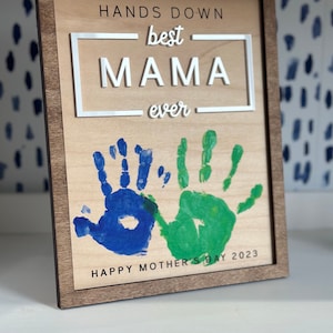 May include: A wooden sign with the text "Hands Down best MAMA ever" and two handprints in blue and green paint. The sign also says "Happy Mother's Day 2023".