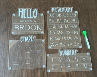May include: Four clear acrylic dry-erase boards with white lettering. The boards are labeled "Hello", "Shapes", "The Alphabet", and "Numbers". The "Hello" board has a space for a name, the "Shapes" board has outlines of different shapes, the "Alphabet" board has the letters of the alphabet, and the "Numbers" board has the numbers 1 through 10.