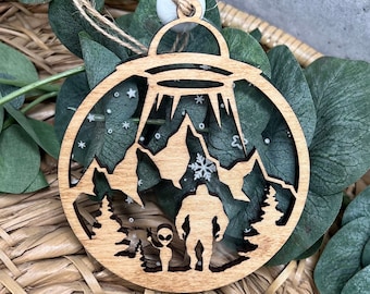 May include: A wooden Christmas ornament shaped like a circle with a UFO, mountains, a Bigfoot, and an alien inside. The ornament is decorated with white glitter and snowflakes.