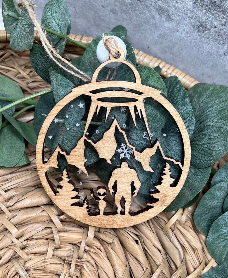May include: A wooden Christmas ornament shaped like a circle with a UFO, mountains, a Bigfoot, and an alien inside. The ornament is decorated with white glitter and snowflakes.