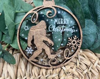 May include: A wooden Christmas ornament shaped like a round ball with a clear center. The ornament features a silhouette of a Bigfoot with the text "Merry Christmas" inside. The ornament is decorated with white snowflakes and stars.
