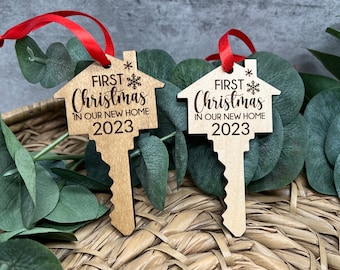 May include: Two wooden key-shaped Christmas ornaments. Each ornament has a house-shaped top with the words "First Christmas in our new home 2023" and a snowflake design. Red ribbons are attached for hanging.