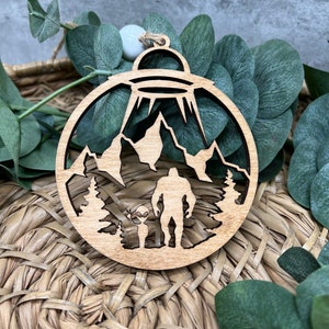 May include: A wooden Christmas ornament featuring a silhouette of a Bigfoot and an alien standing in a forest with mountains in the background. A UFO hovers above them.