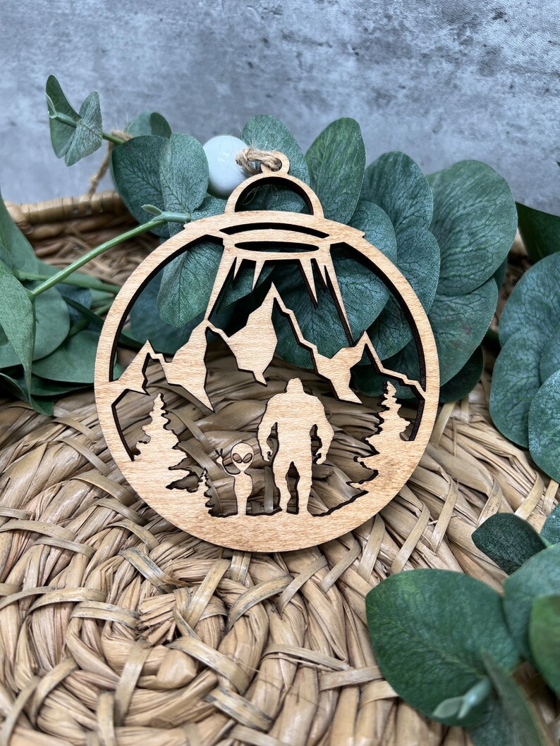 May include: A wooden Christmas ornament featuring a silhouette of a Bigfoot and an alien standing in a forest with mountains in the background. A UFO hovers above them.