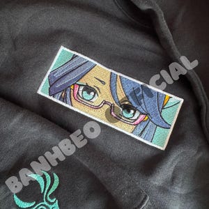 May include: Black hoodie featuring an embroidered patch of an anime character with blue hair and glasses. The patch is framed with a white border and has a light blue background. The text "OFFICIAL" is partially visible on the right side of the patch.