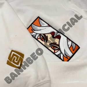 May include: White sweatshirt with an orange and white embroidered image of a character with red eyes and white hair. The text "OFFICIAL" is embroidered in black on the right side of the image. The text "BANHBE0" is embroidered in gold on the left side of the image.