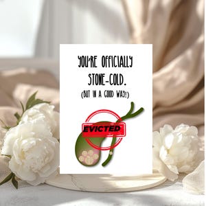 Könnte beinhalten: Eine weiße Grußkarte mit dem Text "YOU'RE OFFICIALLY STONE-COLD. (BUT IN A GOOD WAY!)". Die Karte zeigt eine grüne Abbildung einer Gallenblase mit Steinen und einen roten "EVICTED" Stempel. Weiße Blumen sind im Hintergrund.