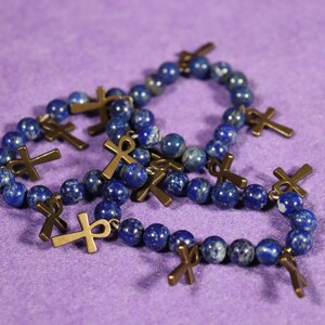 May include: Several bracelets with dark blue, spherical beads and bronze-colored ankh charms are displayed on a purple surface. The ankh charms are attached to the bracelets. The bracelets are likely jewelry.