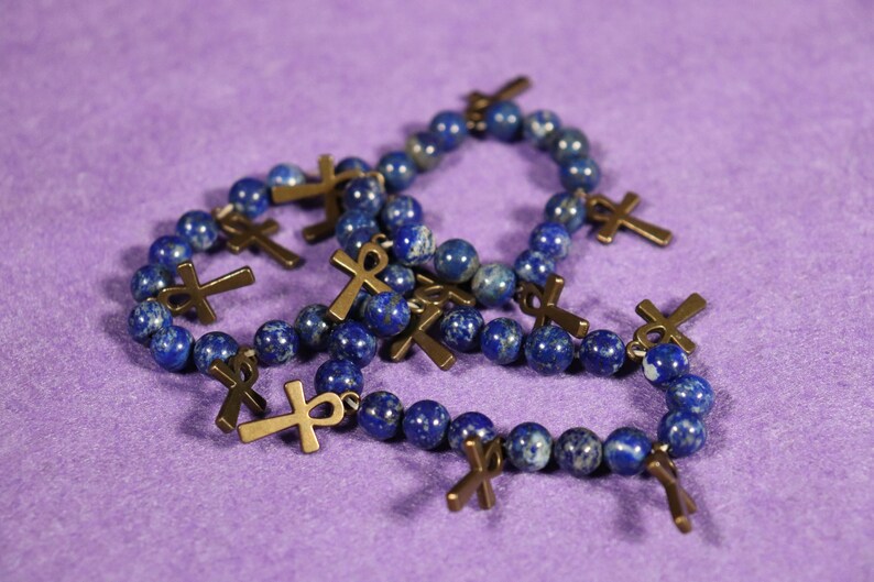 May include: Several bracelets with dark blue, spherical beads and bronze-colored ankh charms are displayed on a purple surface. The ankh charms are attached to the bracelets. The bracelets are likely jewelry.