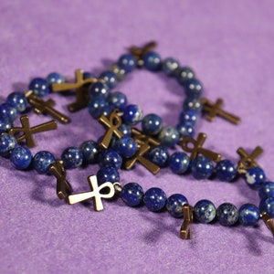 May include: Several beaded bracelets with dark blue, speckled beads and bronze-colored ankh charms are displayed on a purple surface. The bracelets are arranged in a casual, overlapping manner.