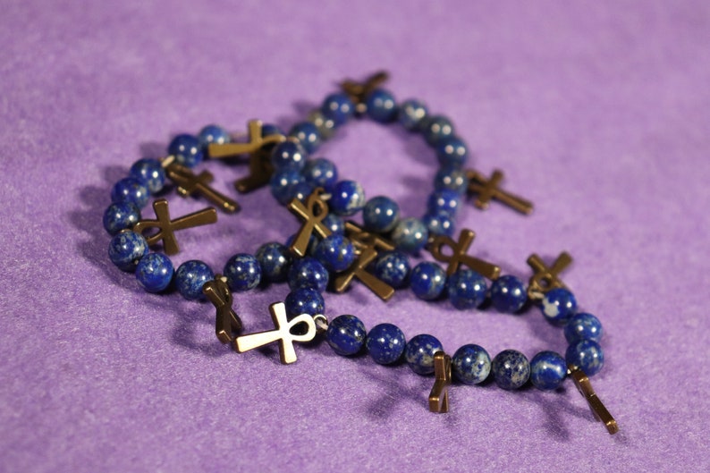 May include: Several beaded bracelets with dark blue, speckled beads and bronze-colored ankh charms are displayed on a purple surface. The bracelets are arranged in a casual, overlapping manner.