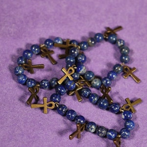 May include: A collection of beaded bracelets with dark blue lapis lazuli beads and bronze-colored ankh and cross charms. The bracelets are arranged on a purple surface, showcasing the jewelry's details.