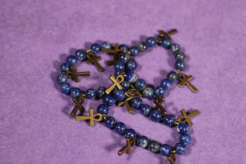 May include: A collection of beaded bracelets with dark blue lapis lazuli beads and bronze-colored ankh and cross charms. The bracelets are arranged on a purple surface, showcasing the jewelry's details.