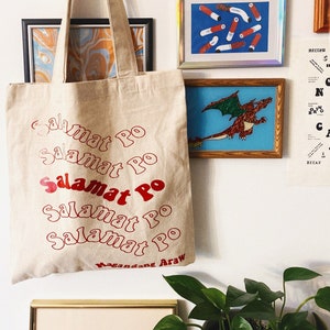 May include: A beige canvas tote bag with red text that says "Salamat Po" multiple times. The words "Magandang Araw" are printed at the bottom. The bag hangs on a white wall with framed artwork and a plant.