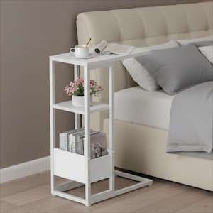 May include: A white, C-shaped side table with a built-in shelf and magazine rack. A cup of coffee, a small potted plant, and books are on the table. The table is next to a bed with a beige headboard and gray pillows.