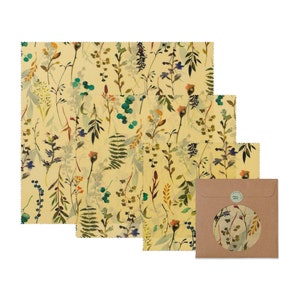 May include: A set of three beeswax food wraps featuring a floral design in shades of green, yellow and brown. The wraps are packaged in a brown paper envelope with a circular window revealing the pattern.