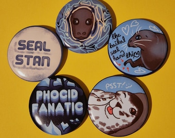 May include: A collection of five round buttons with seal illustrations and text. The buttons feature various designs, including a seal face, a seal with hearts, and the phrases "Seal Stan" and "Phocid Fanatic."