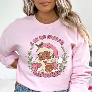 Op de afbeelding: Een lichtroze sweatshirt met een afbeelding van de kerstman die knipoogt, een roze muts en wanten draagt en een mok vasthoudt. De tekst "A MI ME GUSTAN BARBONES" is boven en onder de Santa-illustratie gebogen, omgeven door een groene krans.