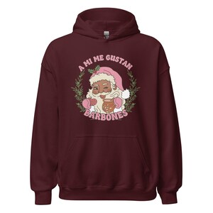 Op de afbeelding: Een bordeauxrode hoodie met een Santa Claus-afbeelding met een roze muts en wanten, die een mok vasthoudt. De afbeelding is omgeven door een krans en de tekst "A MI ME GUSTAN BARBONES".