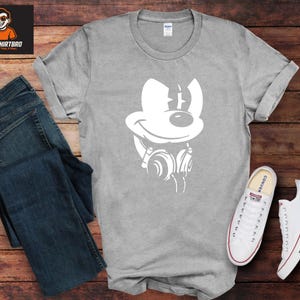 May include: A grey t-shirt featuring a white graphic of a cartoon mouse with headphones. The shirt is displayed with blue jeans and white trainers with red and blue details. The shirt is on a wooden surface.