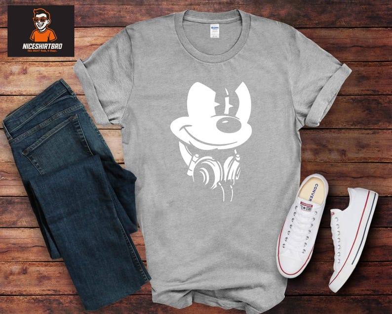 May include: A grey t-shirt featuring a white graphic of a cartoon mouse with headphones. The shirt is displayed with blue jeans and white trainers with red and blue details. The shirt is on a wooden surface.
