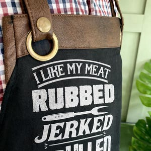May include: Black apron with brown leather straps and the text "I LIKE MY MEAT RUBBED JERKED & PULLED" in white. The apron has a brass-colored metal ring and button detail. The background includes a plaid shirt.