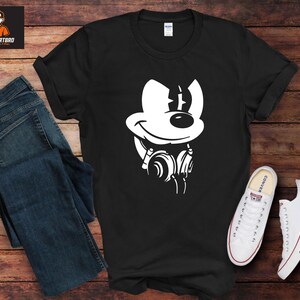 May include: Black t-shirt with a white cartoon mouse graphic wearing headphones. The image includes blue jeans and white trainers. The background is a wooden surface.