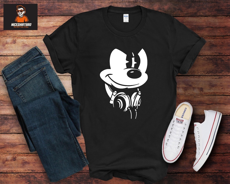 May include: Black t-shirt with a white cartoon mouse graphic wearing headphones. The image includes blue jeans and white trainers. The background is a wooden surface.