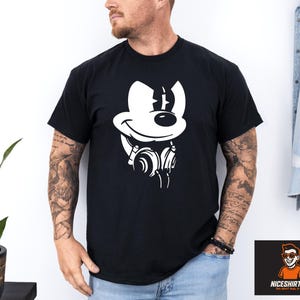 May include: Black t-shirt with a white cartoon mouse graphic wearing headphones. The mouse has a large smile and a line on its head. The shirt is worn by a person with tattoos on their arm.