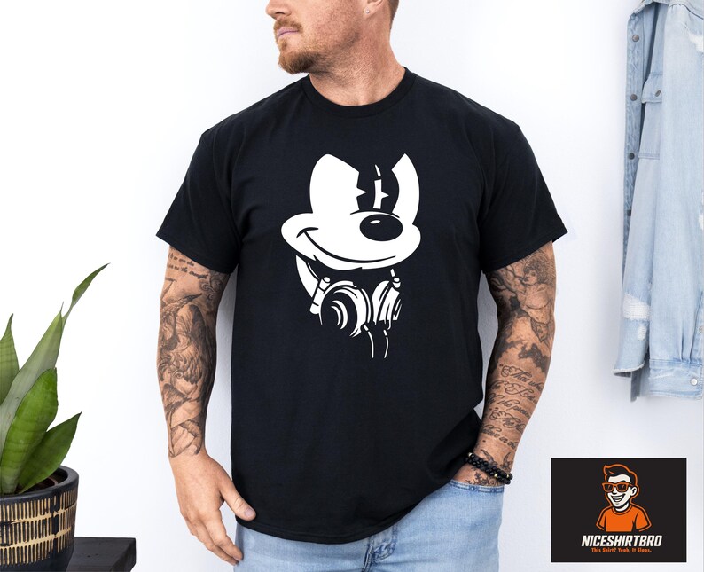 May include: Black t-shirt with a white cartoon mouse graphic wearing headphones. The mouse has a large smile and a line on its head. The shirt is worn by a person with tattoos on their arm.
