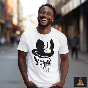 May include: White t-shirt with a black cartoon graphic of a character wearing a hat and headphones. The design includes a smiling face. The shirt is worn by a person smiling outdoors.