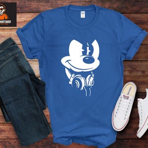May include: A royal blue t-shirt with a white cartoon character design wearing headphones. The image is next to a pair of blue jeans and white trainers with red and blue stripes on the soles. The shirt is on a wooden surface.