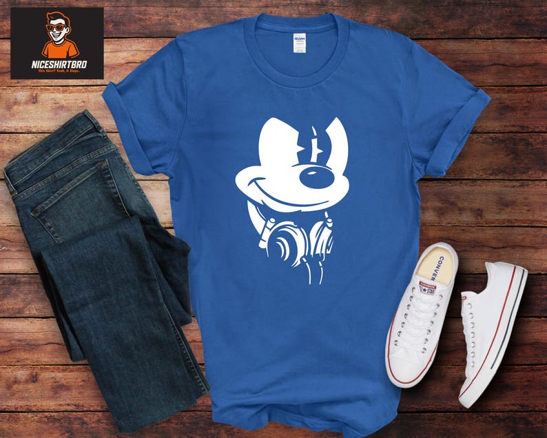 May include: A royal blue t-shirt with a white cartoon character design wearing headphones. The image is next to a pair of blue jeans and white trainers with red and blue stripes on the soles. The shirt is on a wooden surface.