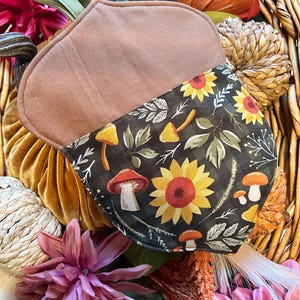 May include: A brown acorn-shaped face mask with a floral pattern of sunflowers and mushrooms. The mask has a brown fabric exterior and a black interior with the pattern. The mask is surrounded by fall-themed decorations.