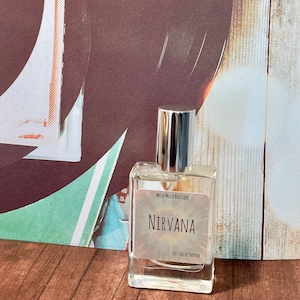 Puede incluir: Un frasco de perfume de vidrio transparente con tapa plateada. La etiqueta dice "Nirvana" y "1 oz. Eau de Parfum". El fondo presenta un diseño abstracto marrón y blanco y una superficie de madera.
