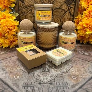 Può includere: Un assortimento di candele e cere profumate a tema autunnale. Le candele sono in barattoli di vetro con coperchi in sughero o legno. La parola "Autumn" è visibile sulle etichette. Sullo sfondo, una scatola di legno decorativa, con fiori arancioni.