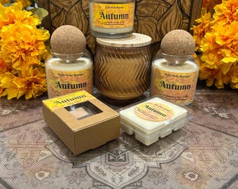 May include: An assortment of autumn-themed candles and wax melts. The candles are in glass jars with cork or wooden lids. The text "Autumn" is visible on the labels. A decorative wooden box is in the background, along with orange flowers.